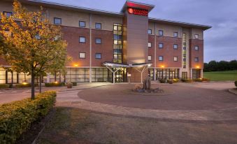 a modern hotel with a large entrance and a red sign on the roof , surrounded by trees and benches at Ramada Plaza by Wyndham Wrexham