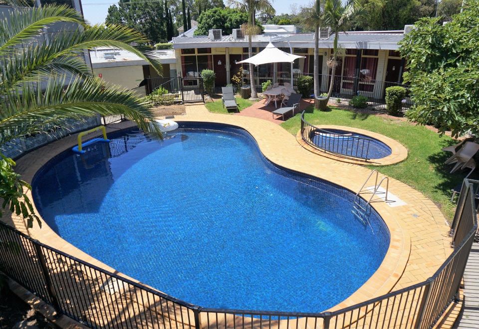 a large , curved swimming pool surrounded by lush greenery and buildings , with umbrellas and chairs nearby at Jacksons Motor Inn