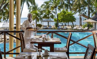 a man in a white robe is standing at an outdoor table with tea and snacks , overlooking a pool and palm trees at Diamonds Dream of Africa