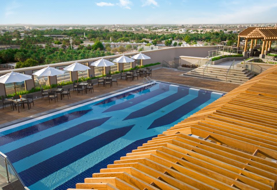 a large swimming pool with a wooden roof is surrounded by umbrellas and has a view of the city at Ayla Grand Hotel Al Ain