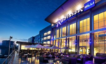 a large building with a restaurant called radisson blu , surrounded by tables and chairs , umbrellas , and people enjoying the outdoor seating area at Radisson Blu Anchorage Hotel