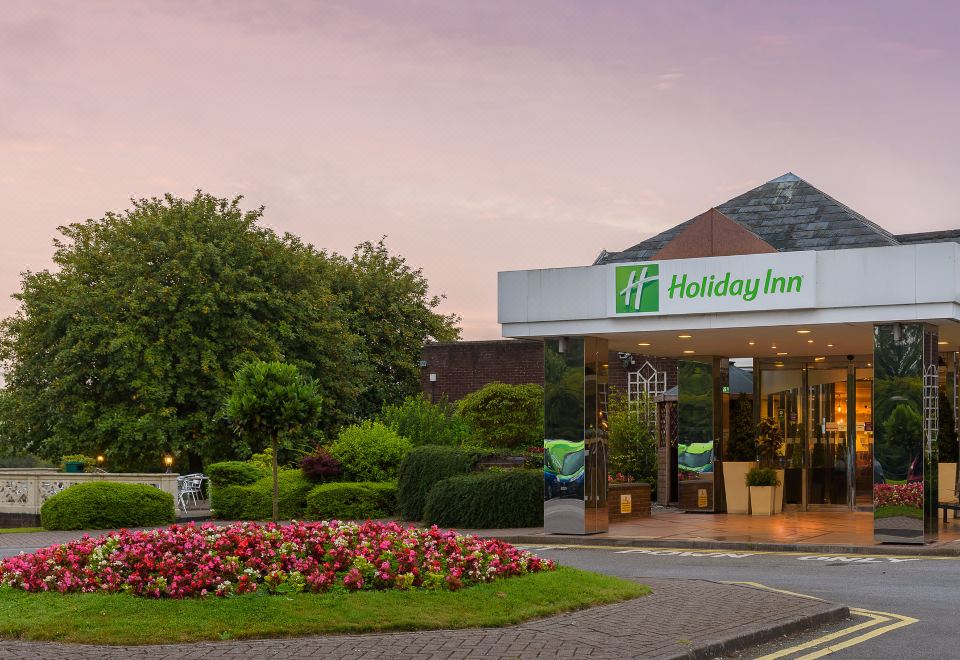 a holiday inn building with a large sign above the entrance , surrounded by flowers and trees at Holiday Inn Leeds - Garforth