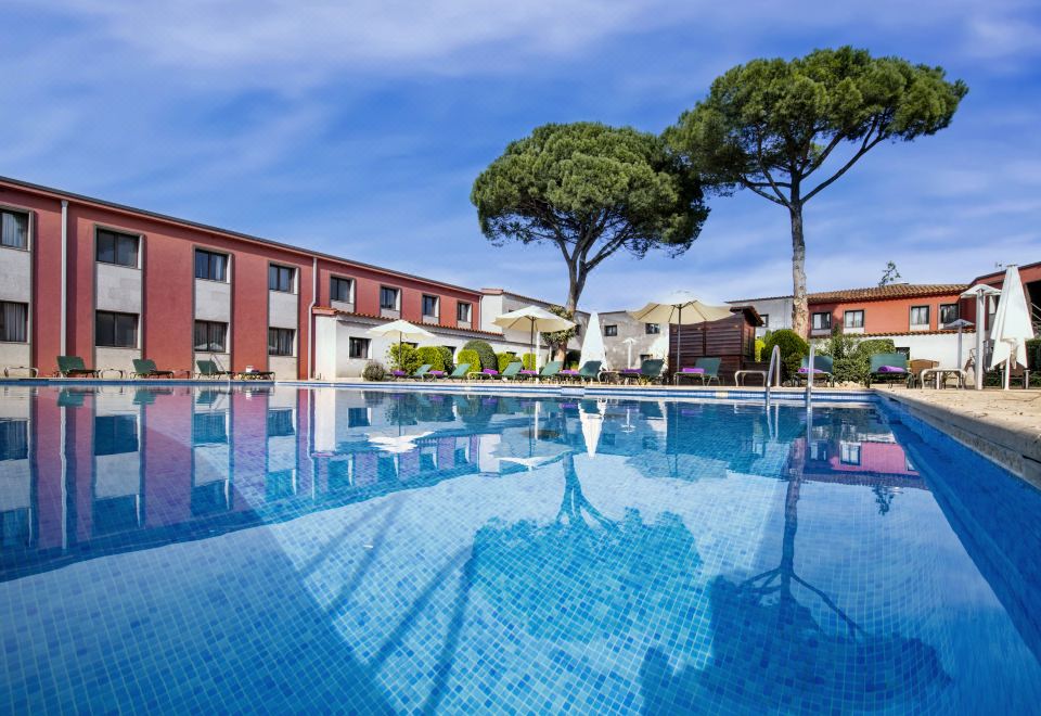 a large outdoor swimming pool surrounded by a building , with trees and umbrellas placed around the pool area at Salles Hotel Aeroport de Girona