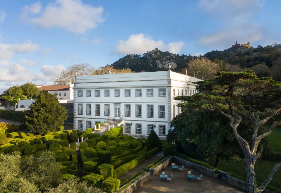 a large white building surrounded by greenery , with a pool and mountains in the background at Valverde Sintra Palacio de Seteais