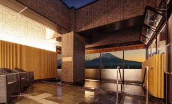 a modern building with a large glass window and stone walls , overlooking a mountainous landscape at dusk at HOTEL MYSTAYS Fuji Onsen Resort