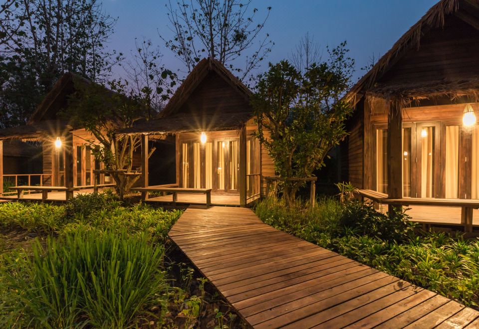 a wooden walkway leads to a row of small , wooden cabins surrounded by lush greenery at night at Azalea Village