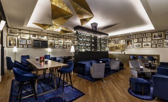 a modern bar with blue chairs , wooden tables , and gold chandeliers , surrounded by framed pictures on the walls at The Oberoi Grand Kolkata