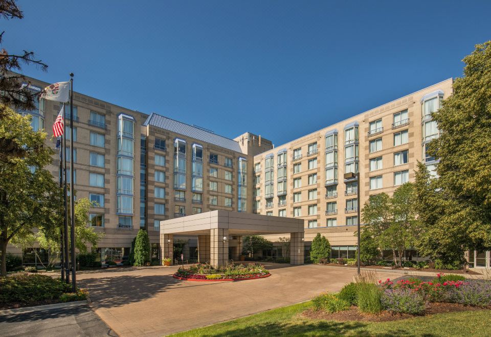 a large hotel with multiple floors , surrounded by trees and flowers , under a clear blue sky at Sheraton Suites Chicago Elk Grove