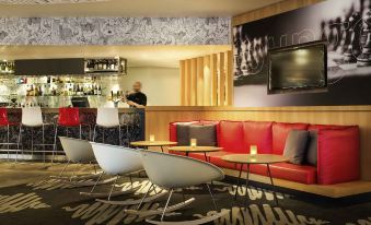 a modern lounge area with red couches , white chairs , and a bar , along with a bartender in the background at Ibis Sydney Airport