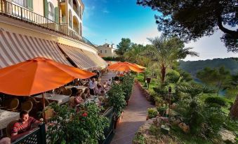 a picturesque outdoor dining area with umbrellas , tables , and chairs , surrounded by lush greenery and a beautiful view of a building at Aparthotel Ona Cala Pi Club