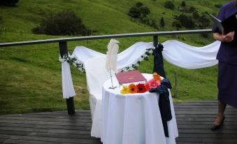 a table with a white tablecloth and flowers is set up on a wooden deck overlooking green hills at Top of the Hill