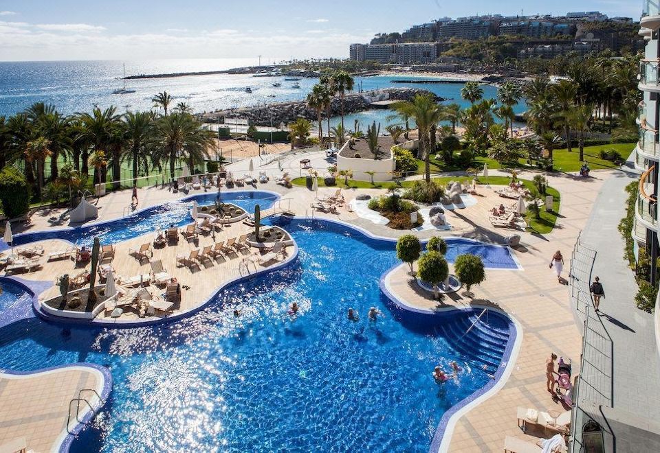 a large swimming pool with a beach and palm trees in the background , surrounded by people enjoying the water at Radisson Blu Resort, Gran Canaria