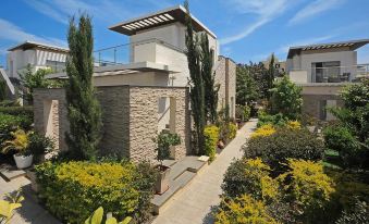 a modern building with a stone wall and white balconies , surrounded by lush greenery and trees at E-Hotel Larnaca Resort & Spa