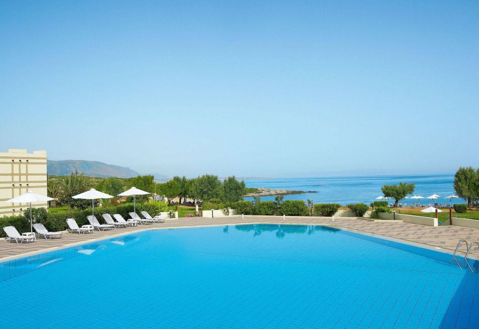 a large outdoor swimming pool surrounded by chairs and umbrellas , with a view of the ocean in the background at Grecotel Meli Palace