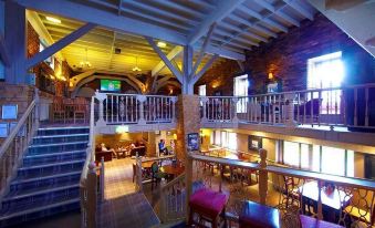 a spacious room with wooden beams and a staircase , featuring tables and chairs , chairs , and a person standing near the window at Village Hotel Manchester Hyde