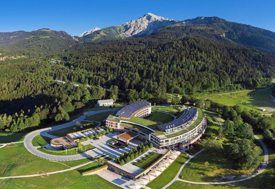 an aerial view of a large hotel surrounded by mountains and greenery , with the sun setting in the background at Kempinski Hotel Berchtesgaden