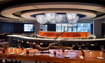 a modern restaurant with a large wooden dining table surrounded by chairs , and a bar area in the background at Doubletree by Hilton Montreal