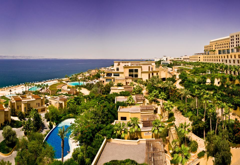a resort with a pool surrounded by lush greenery and the ocean in the background at Kempinski Hotel Ishtar Dead Sea