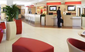 a modern hotel lobby with red and white furniture , people standing near reception desks , and an employee working behind the counter at Ibis Lagos Ikeja