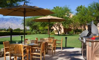 a dining area with wooden chairs and tables under umbrellas , surrounded by a lush green lawn and trees at The Westin Rancho Mirage Golf Resort & Spa