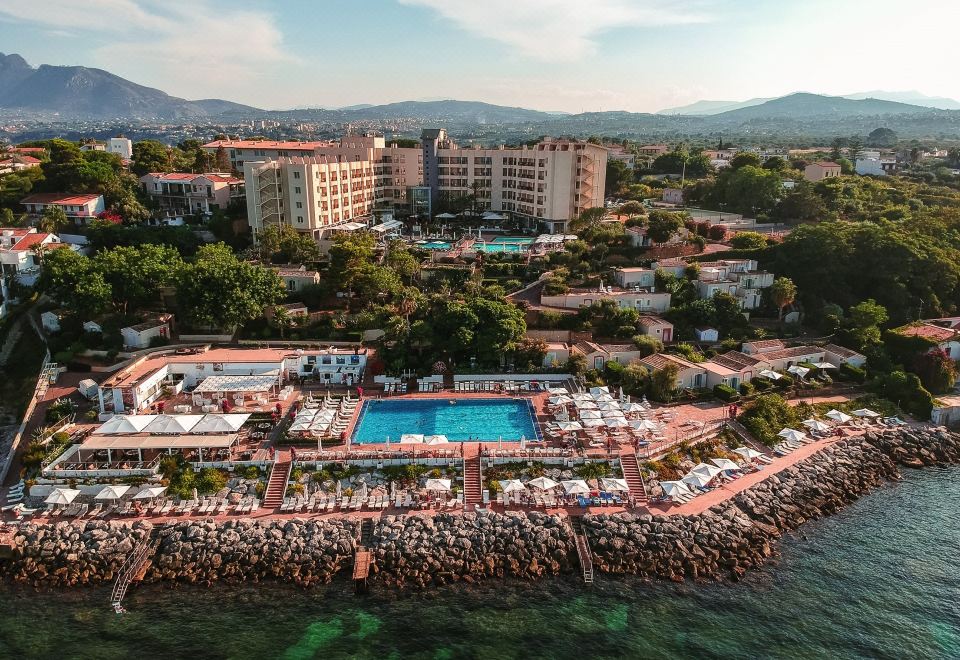 aerial view of a resort with a pool surrounded by palm trees and buildings , situated on the coast near a body of water at Domina Zagarella Sicily