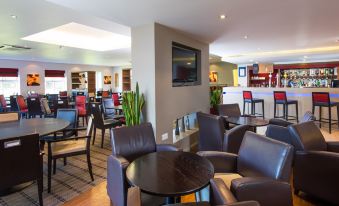 a modern lounge area with black leather chairs , wooden tables , and a large screen tv at Holiday Inn Express Warwick - Stratford-Upon-Avon