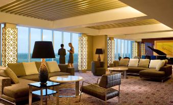 a large , well - lit room with a couch , tables , and chairs is shown with people standing near the windows at Novotel Bangka - Hotel & Convention Centre