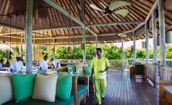 a man wearing a yellow shirt is walking on a wooden deck near a dining area at Six Senses Laamu