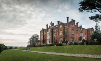 a large , red brick building with multiple chimneys and green grass surrounding it , set against a cloudy sky at De Vere Latimer Estate
