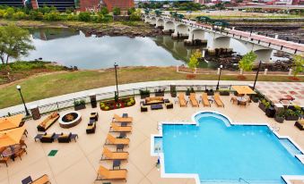 an outdoor pool surrounded by lounge chairs and umbrellas , with a view of a river and a bridge in the background at Courtyard Columbus Phenix City/Riverfront