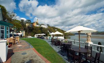 a scenic view of a terrace with umbrellas , tables , and people , overlooking a river and buildings under a cloudy sky at Portmeirion Village & Castell Deudraeth