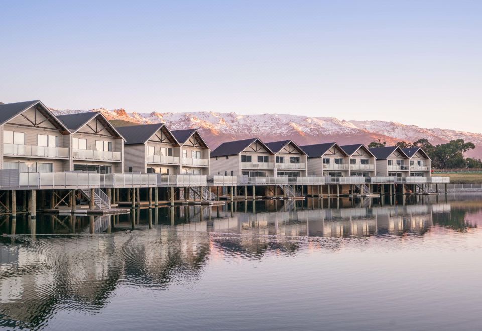 a row of wooden houses is situated on the shore of a lake , with mountains in the background at Marsden Lake Resort Central Otago