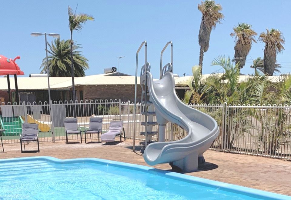 a swimming pool with a silver water slide , surrounded by lounge chairs and palm trees at Carnarvon Motel