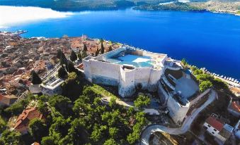 a large white building with a blue swimming pool is perched on the edge of a cliff overlooking a serene lake at Hotel Panorama