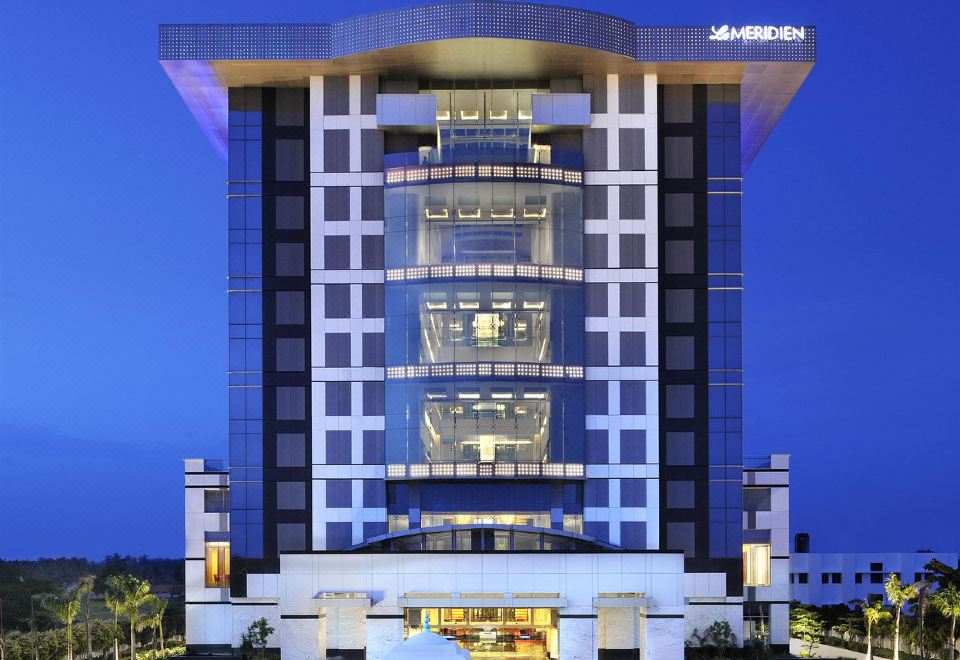 a modern hotel building with a large glass facade and multiple floors , surrounded by palm trees at night at Le Meridien Coimbatore