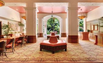 The lobby features chairs and tables in the center, adjacent to the entrance that leads to the hotel's reception area at The Royal Hawaiian, A Luxury Collection Resort, Waikiki