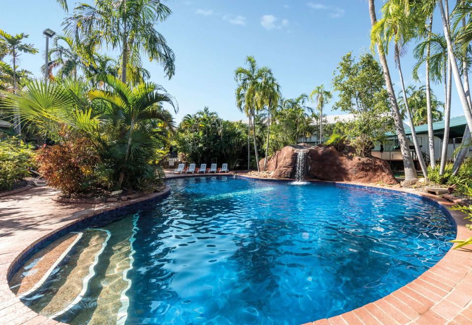 a large swimming pool with a waterfall and lounge chairs is surrounded by palm trees at Travelodge Resort Darwin