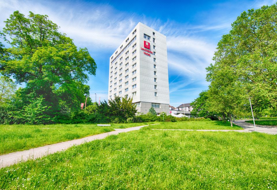 "a tall white building with the name "" petrochach "" on it is surrounded by green grass and trees" at Leonardo Hotel Karlsruhe