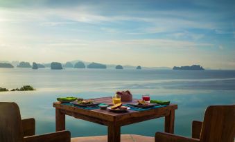 a wooden dining table set up with plates , cups , and utensils in front of a scenic view of water at Six Senses YAO Noi