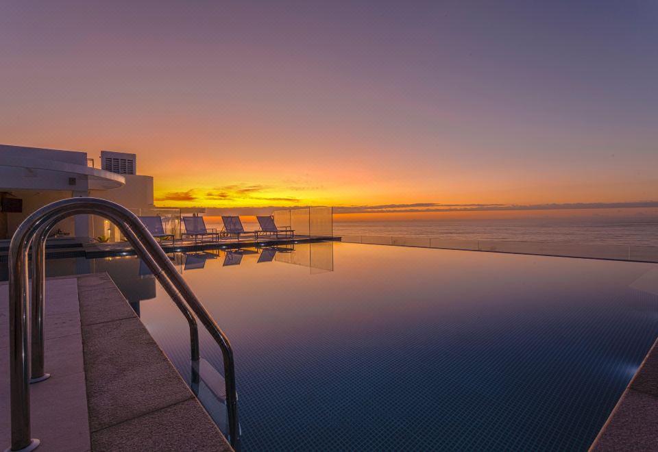 a large infinity pool overlooks the ocean at sunset , with several lounge chairs and umbrellas on the deck at CDesign Hotel