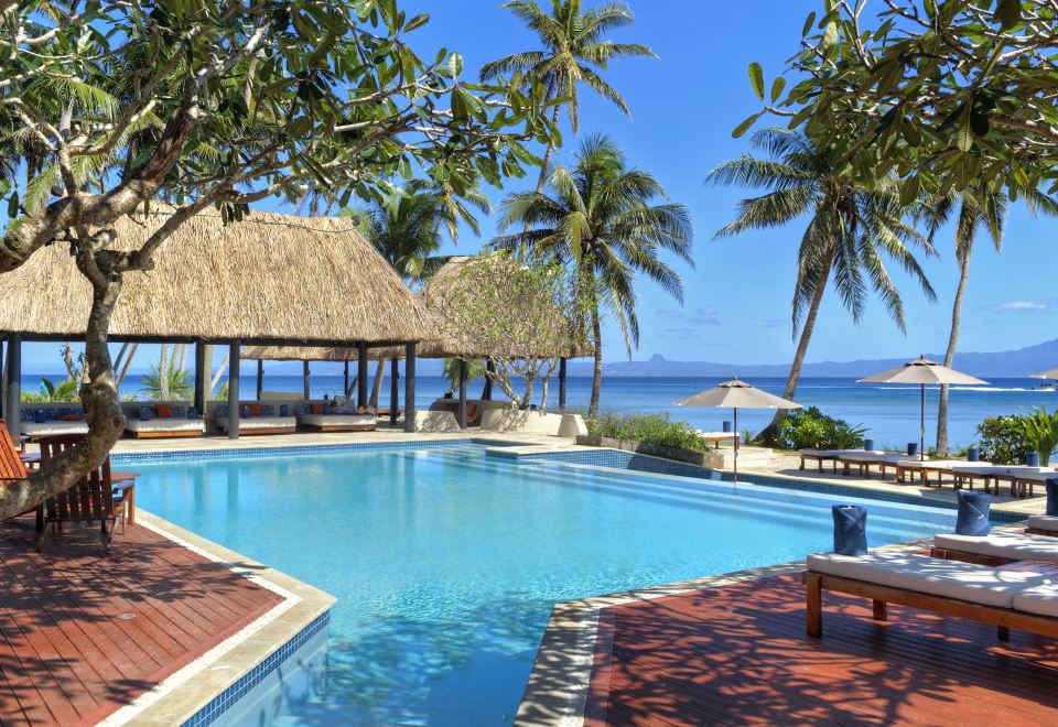 a large swimming pool with a thatched roof hut and palm trees in the background at Jean-Michel Cousteau Resort Fiji