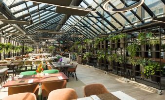 a large dining room with wooden tables and chairs , surrounded by potted plants and a glass roof at NH Amsterdam Schiphol Airport