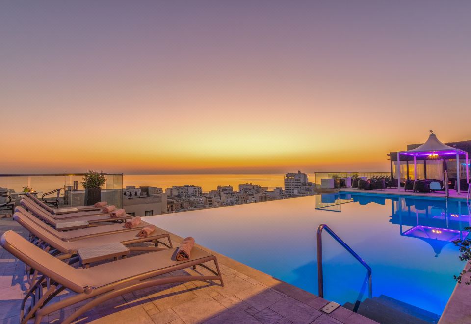 a large swimming pool surrounded by lounge chairs , with a beautiful sunset in the background at AX The Palace