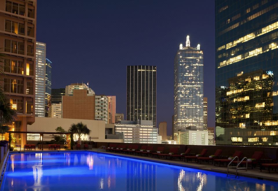 a city skyline at night , with a swimming pool illuminated by blue lights and surrounded by tall buildings at Fairmont Dallas