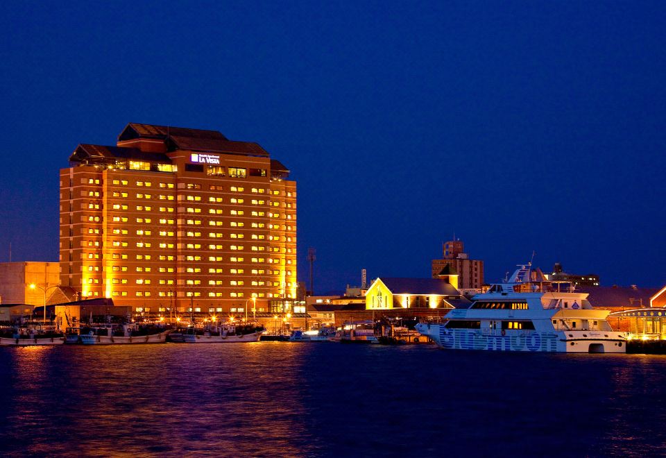 a large hotel building surrounded by a marina at night , with boats docked nearby and lights illuminating the scene at La Vista Hakodate Bay