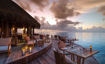 a beachside restaurant with wooden tables and chairs , umbrellas , and lanterns , as the sun sets over the ocean at Mirihi Island Resort