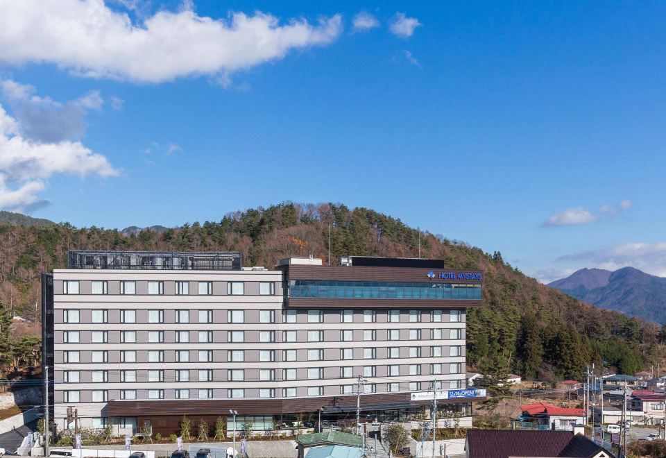 a large hotel with a blue roof is surrounded by trees and mountains , under a clear blue sky at HOTEL MYSTAYS Fuji Onsen Resort