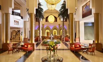 a luxurious hotel lobby with high ceilings , marble floors , and an elegant chandelier , surrounded by red sofas and tables at Ajman Hotel by Blazon Hotels