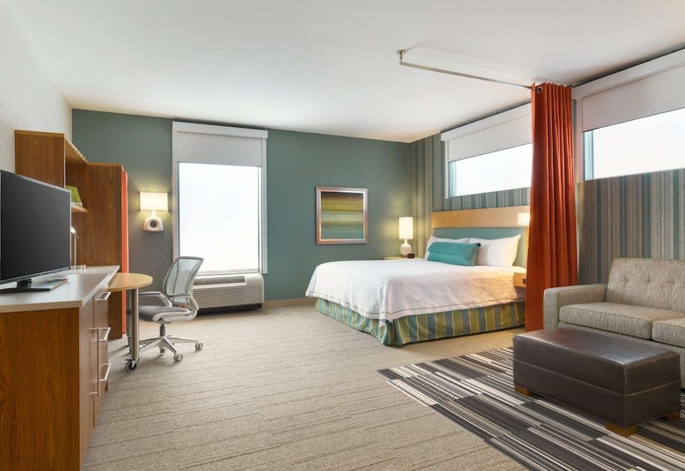 a modern hotel room with wooden flooring , blue walls , and white bedding , along with a desk , chair , and large window at Home2 Suites by Hilton Downingtown Exton Route 30