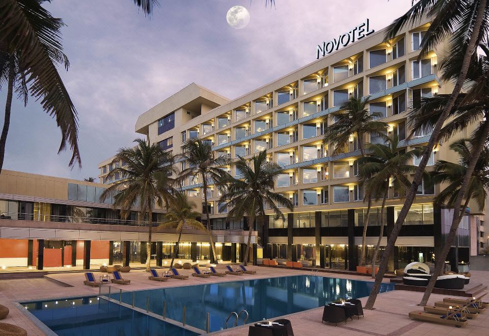a large hotel with a pool and palm trees is shown during the night , with a full moon in the background at Novotel Mumbai Juhu Beach